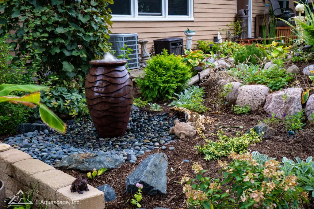 A scalloped urn fountain nestled in a garden mulch bed, gently spilling water over its ribbed edges. It's surrounded by a bed of smooth river rocks and an assortment of lush plants, including ferns and flowering shrubs. This naturalistic setting is complemented by larger boulders and a variety of foliage, creating an inviting habitat for local wildlife and adding a soothing water element to the landscape. The scene is set against a home with neutral siding, integrating the water feature into the residential outdoor space.