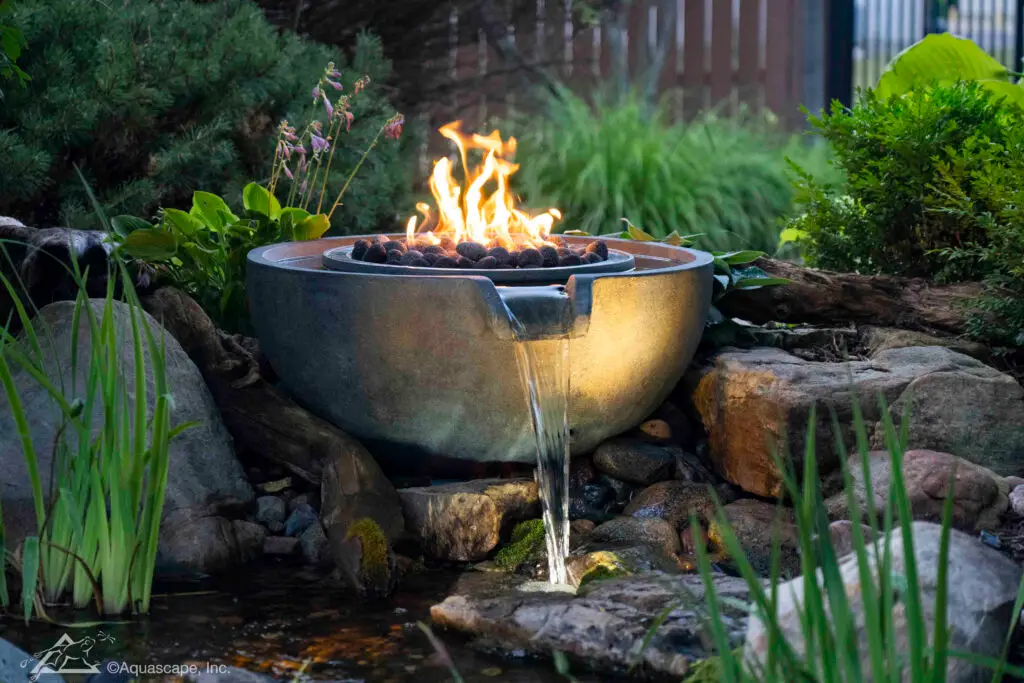 A captivating spillway bowl with a central fire feature creates a striking balance of elements in a garden setting. The fire's warm glow reflects off the water cascading from the bowl into a rocky stream below. Surrounded by rich foliage and soft evening light, this fusion of fire and water provides a mesmerizing focal point and adds a sense of mystique and elegance to the outdoor space.