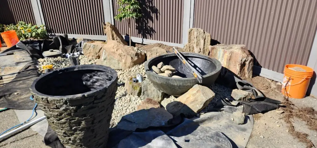 A work-in-progress installation of a water feature, with a slate stacked urn and bowl visible amidst construction materials. The area is being prepared with an underlayment and large rocks, suggesting the feature’s integration into a landscape. An orange bucket, level, and loose soil are scattered around, indicating ongoing work. The backdrop shows part of a building and a young tree, providing context for the future site of the water feature.