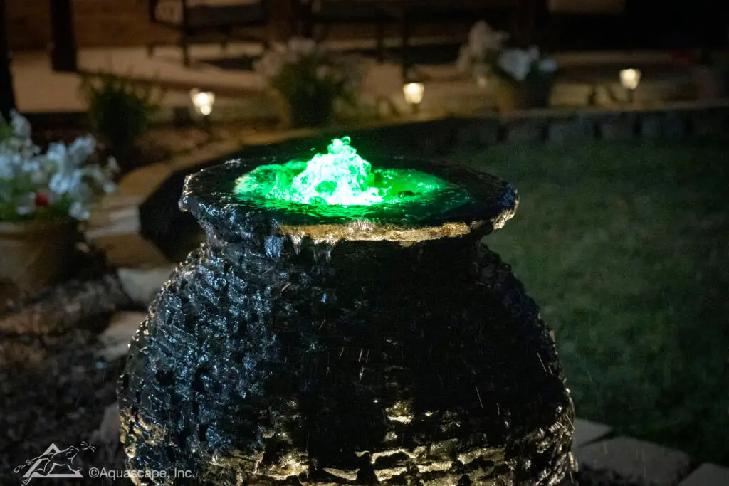 A nighttime view of a slate stacked sphere fountain illuminated by a vibrant green light, creating a mesmerizing water display. The light accentuates the water's gentle cascade over the textured stone surface, while the surrounding darkness adds to the fountain's dramatic effect. The soft ambient lighting in the garden hints at the presence of surrounding plants, creating a tranquil and enchanting atmosphere in the outdoor space.