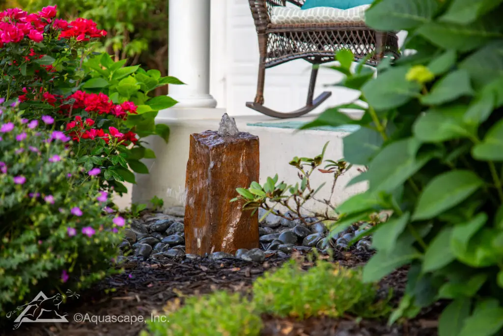 A faux basalt column fountain creates a subtle yet striking water feature in a garden setting, with water gently spouting at the top. Surrounded by polished river stones and set against a backdrop of vibrant flowering plants and lush foliage, the feature adds a naturalistic touch to the landscape. The comfortable porch seating in the background invites relaxation and enjoyment of the tranquil sounds of flowing water in this peaceful garden nook.