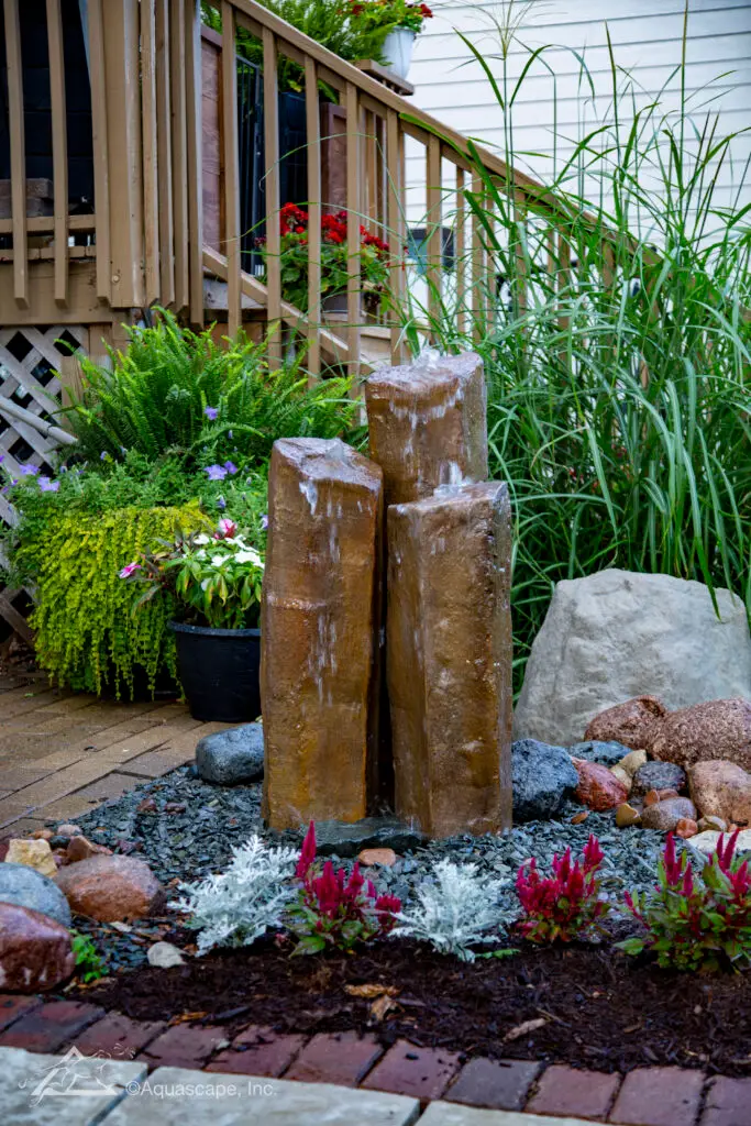 A set of three faux basalt column fountains of varying heights, with water gently cascading down their sides. They're surrounded by a diverse arrangement of garden plants, textured ground cover, and multicolored stones, integrating naturally into the landscaped area. Positioned on a brick patio adjacent to a wooden deck railing, these columns enhance the garden's aesthetic and add a soothing auditory element to the space.