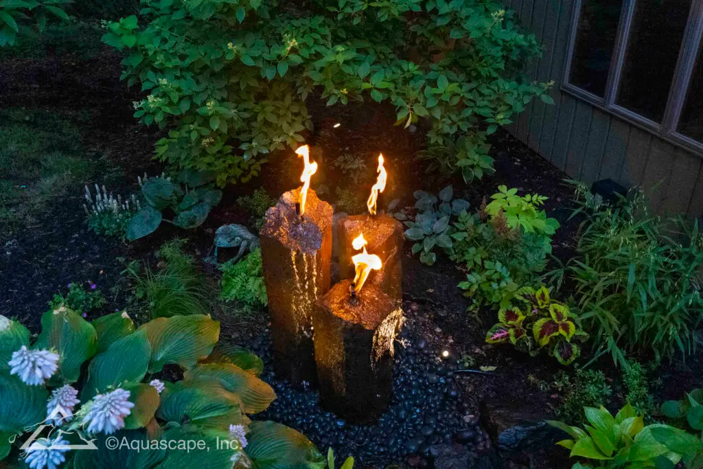 A serene evening scene is brought to life with a trio of fire-fountain basalt columns, each with a robust flame gently flickering at its top. Water cascades down the rugged, textured sides of the basalt into a bed of smooth, dark pebbles. The fire's warm hues contrast with the cool dusk, while the surrounding vegetation—lush hostas, delicate flowers, and a variety of ground cover—adds to the tranquil and natural atmosphere of the garden.