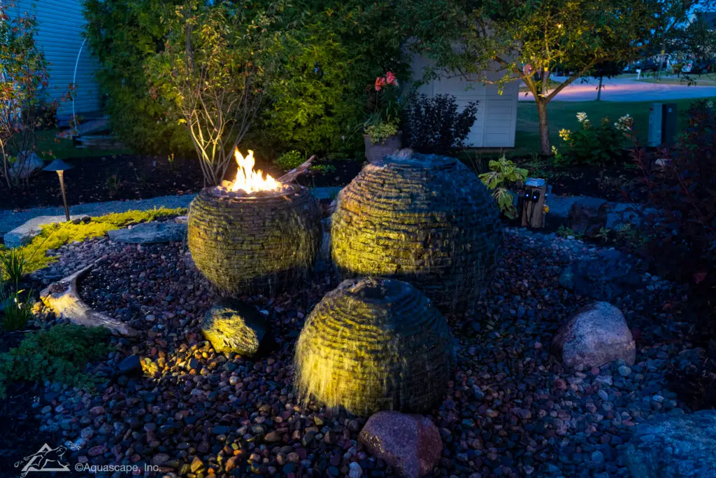 An enchanting evening view of a water feature with three stacked slate sphere fountains of varying sizes. The largest sphere is crowned with a captivating fire feature that illuminates the surrounding area with a warm glow. The fountains are positioned amidst a bed of multicolored pebbles, accompanied by strategically placed boulders and a diverse array of foliage that enhances the naturalistic ambiance of the space.