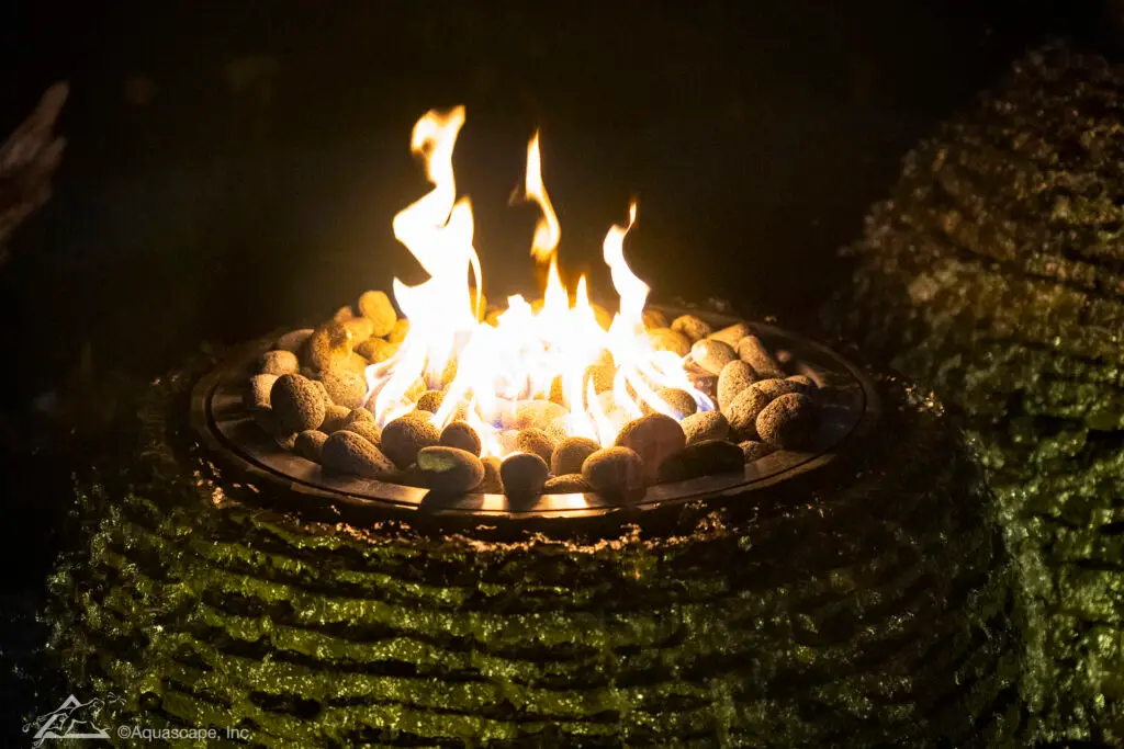 A close-up of a fire feature atop a stacked slate sphere fountain at night. Flames dance among the lava rocks within the burnished metal ring, providing a warm, flickering light that contrasts with the dark, textured stone. This fusion of fire and water elements creates a dramatic visual effect, adding an inviting ambiance to the evening garden setting.