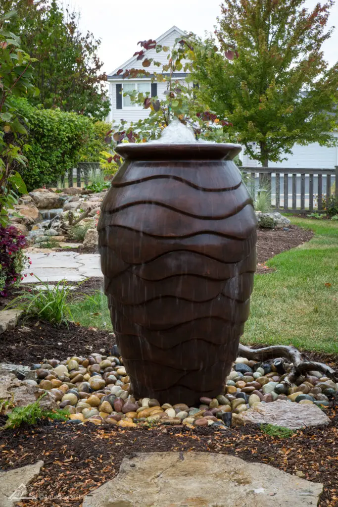 A large scalloped urn fountain sits as the centerpiece in a garden, with water gently bubbling over the top and flowing down its sculpted sides. The urn's dark patina finish contrasts with the surrounding layer of multi-colored river rocks. A flagstone pathway meanders in the background, leading to a landscaped yard with a white picket fence and a traditional white home, contributing to a classic and elegant outdoor ambiance.