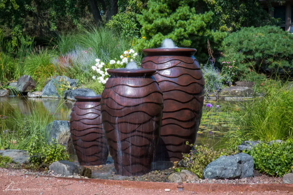 A trio of scalloped urn fountains arranged in a staggered formation, each spilling water smoothly from their brims. The richly toned urns are set against a garden backdrop with a pond, rocks, and a variety of plants, contributing to a serene and naturalistic landscape. The calming presence of the water and the organic arrangement of the urns create a harmonious outdoor feature.