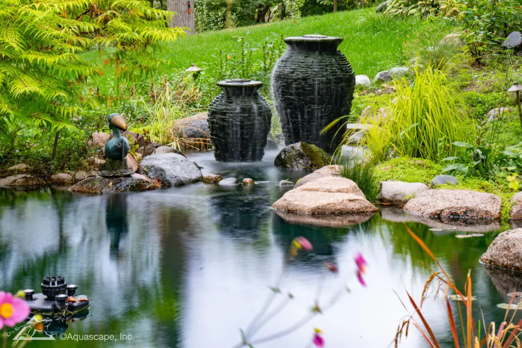 A serene pond featuring two urn fountains with water cascading gently into the pond, creating soft ripples on the water's surface. The setting is tranquil, surrounded by lush greenery, rocks, and blooming plants, with the reflection of the urns and vegetation mirrored in the still water.