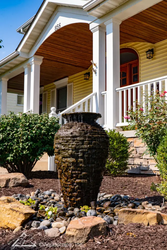 A stately slate stacked urn fountain prominently positioned in the front yard of a home, creating a welcoming water feature. The urn’s textured surface is highlighted by the flow of water, surrounded by a natural arrangement of smooth river rocks and accented with large stones and mulch. Lush shrubs and a few flowers add greenery to the setting, with the home's classic porch, white columns, and warm wood ceiling in the background, offering a picturesque curb appeal.