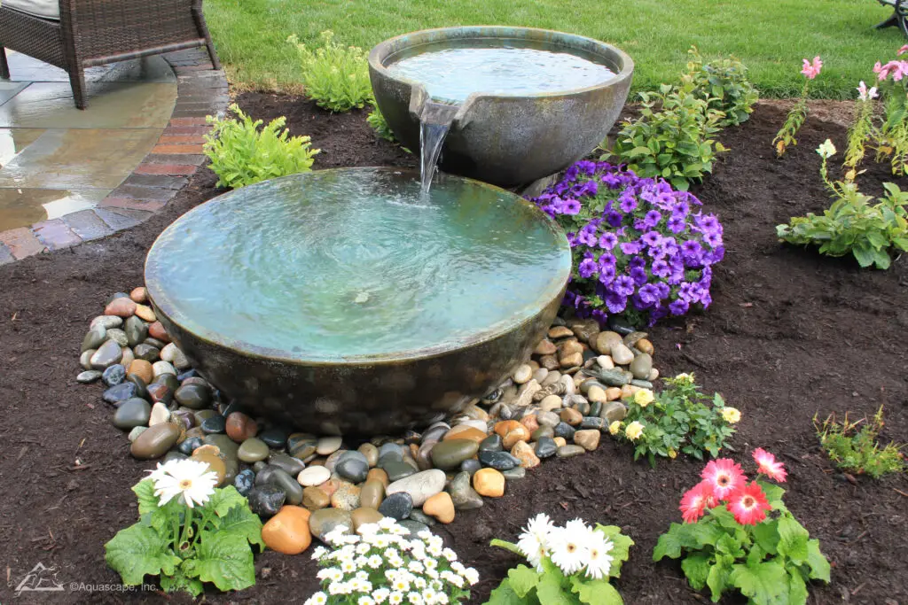 A captivating garden scene showcasing a spillway bowl and basin setup. The spillway bowl at the top overflows into a large basin beneath it, which in turn spills over into an unseen reservoir, creating a continuous and serene water cycle. The arrangement is set amongst a rich array of garden flora, including vivid purple petunias and cheerful daisies, with a backdrop of dark, nourishing mulch that accentuates the lush greenery and floral colors.