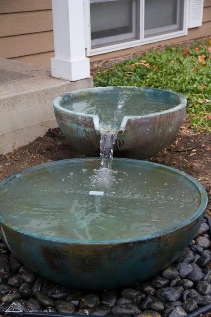 A two-tiered spillway bowl fountain, with water flowing from the smaller upper bowl into the larger basin below. The fountain features a patinated finish and is nestled on a bed of polished river rocks, enhancing the naturalistic setting. Adjacent to the home’s exterior, this fountain provides a focal point in the garden, inviting a sense of peace with the gentle sound of moving water.