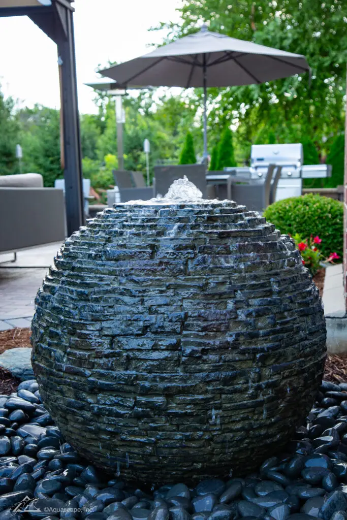 A stacked slate sphere fountain sits as a striking centerpiece on a patio, with water gently cascading over its textured layers. Nestled among polished black pebbles, it complements the outdoor living area, which includes comfortable lounge chairs under a large umbrella and a grill in the background. The arrangement offers an ideal setting for relaxation and entertainment, blending the soothing presence of water with the comforts of a well-appointed backyard retreat.