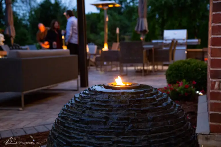 An evening setting showcases a slate stacked sphere with a small, open flame on top, providing a warm and inviting light. The sphere is situated on a patio corner near brickwork, with blurred background activity of people mingling and a well-equipped outdoor kitchen. The ambiance is cozy and social, with the fire feature adding a touch of elegance and comfort to the outdoor entertainment space.