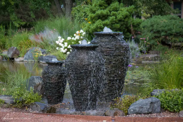 A trio of textured stacked slate urns, nested amidst a lush garden setting, serves as a captivating focal point. The largest urn stands prominently in the center, with a smaller urn on each side, all with water gracefully overflowing their rims. The backdrop features a mix of vibrant white and soft purple flowers, flourishing grasses, and a tranquil pond, enhancing the naturalistic appeal of the scene.