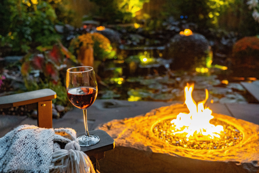 Cozy nighttime outdoor scene featuring a glowing stone fire pit with flames dancing, a glass of red wine resting on an armchair alongside a warm knit blanket. In the background, a softly lit pond and lush greenery create a tranquil, inviting ambiance.