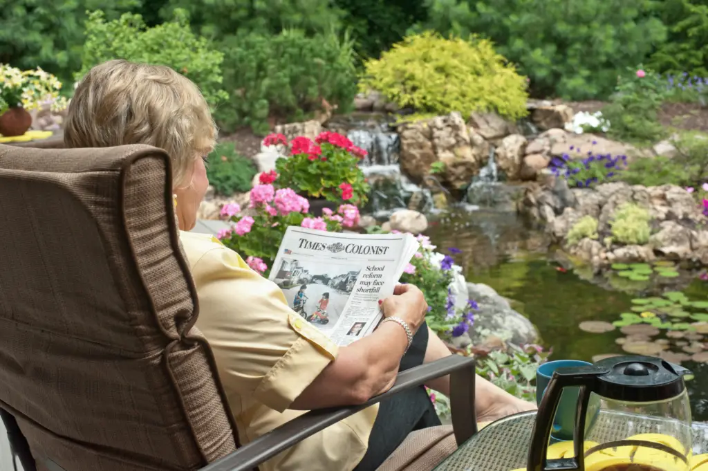 A lady is sitting beside her pond, you can see her coffee pot and newspaper over her shoulder