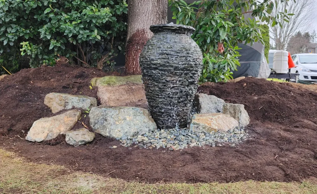 A newly installed urn fountain feature stands as a centerpiece in a mound of rich, dark soil, ready for planting. The urn is textured with a natural stone appearance, complemented by a scattering of river rocks at its base and larger stones partially embedded around the mound. Water gently cascades over the surface of the urn into a hidden basin below. The work-in-progress landscape scene hints at a transformation, integrating the water feature into a suburban front yard with a vehicle and tools indicating ongoing garden work.