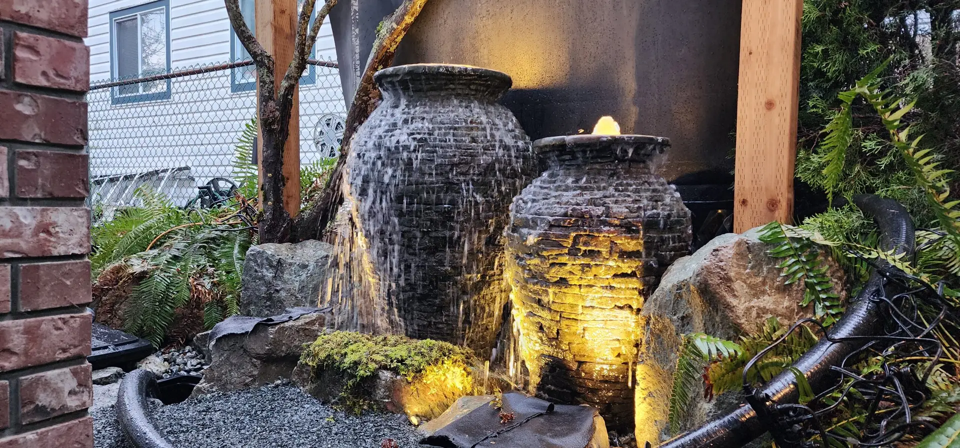 This image shows a garden setup with a large urn and a smaller one, both with water spilling over their edges, set among a rich arrangement of rocks, ferns, and other lush greenery. A light inside the smaller urn creates a captivating glow that illuminates the water and surrounding plants, creating a serene and enchanting ambiance in the outdoor space. The garden is a blend of natural textures and elements, skillfully arranged to create a harmonious and inviting water feature.