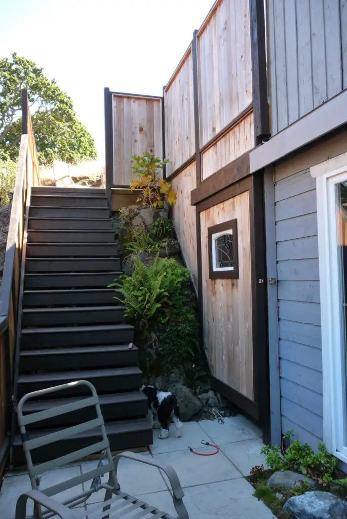 Completed project showing newly installed wooden deck stairs with railings, custom sliding door with a stained-glass window for under-deck storage, and greenery planted along the rock face. The boulder step and plants soften the area, enhancing the overall look and safety.