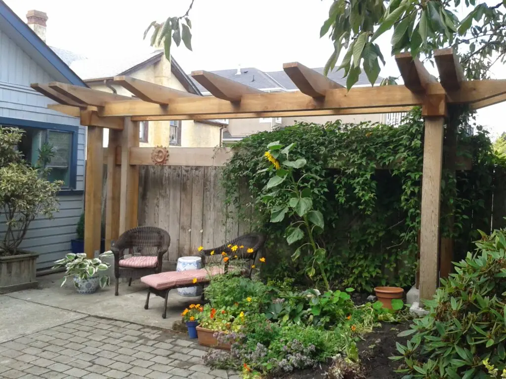 This image shows a cozy outdoor seating area underneath a wooden timber arbor in a small garden. The arbor features sturdy beams with a rustic design, blending naturally into the garden's aesthetic. Two wicker chairs, one with a cushioned footrest, are arranged near a weathered wooden fence. The fence is adorned with a climbing plant that creates lush greenery and partial privacy. A sunflower and various other plants add vibrant color and a natural, inviting feel to the scene. The paved stone patio contrasts with the soft foliage, creating a comfortable, tranquil space for relaxation.