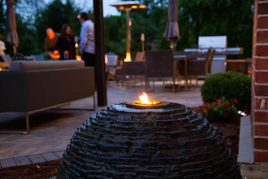 Evening scene of a patio area with a stacked slate sphere fountain topped with a small fire feature in the foreground. In the background, people are gathered near outdoor furniture and a grill, enjoying a cozy atmosphere with additional lighting from patio heaters and candles. The setting is surrounded by trees and shrubs, creating a warm and inviting outdoor space.