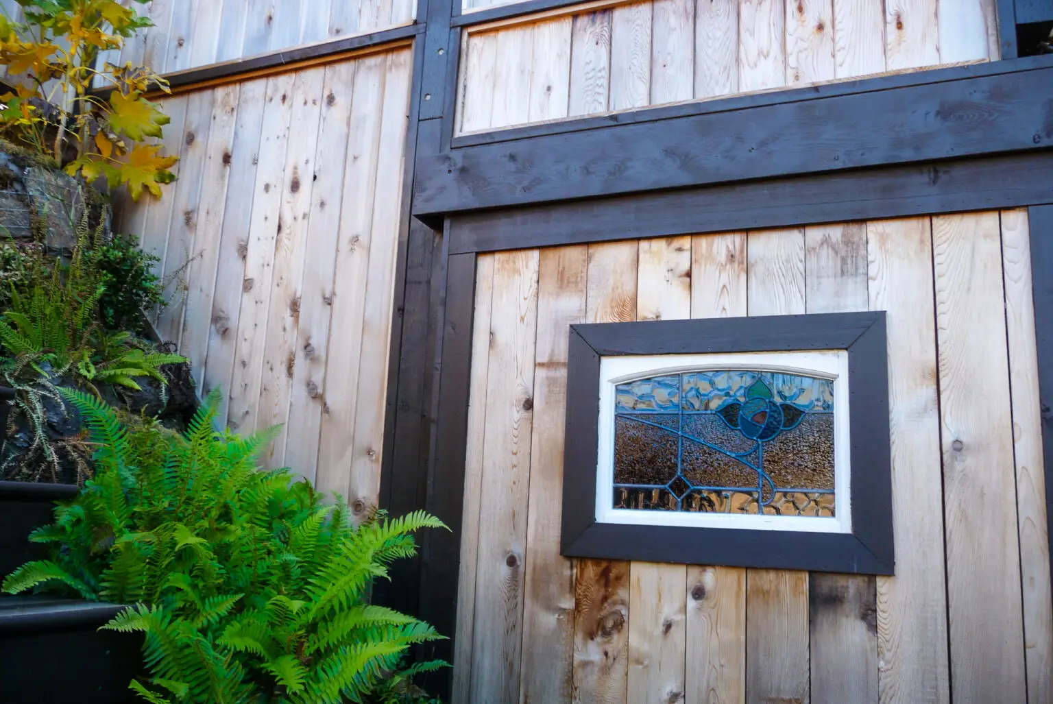 Close-up of a custom sliding door with a stained-glass window, part of under-deck storage. Ferns and greenery grow next to the door, softening the surrounding rock and wood elements.