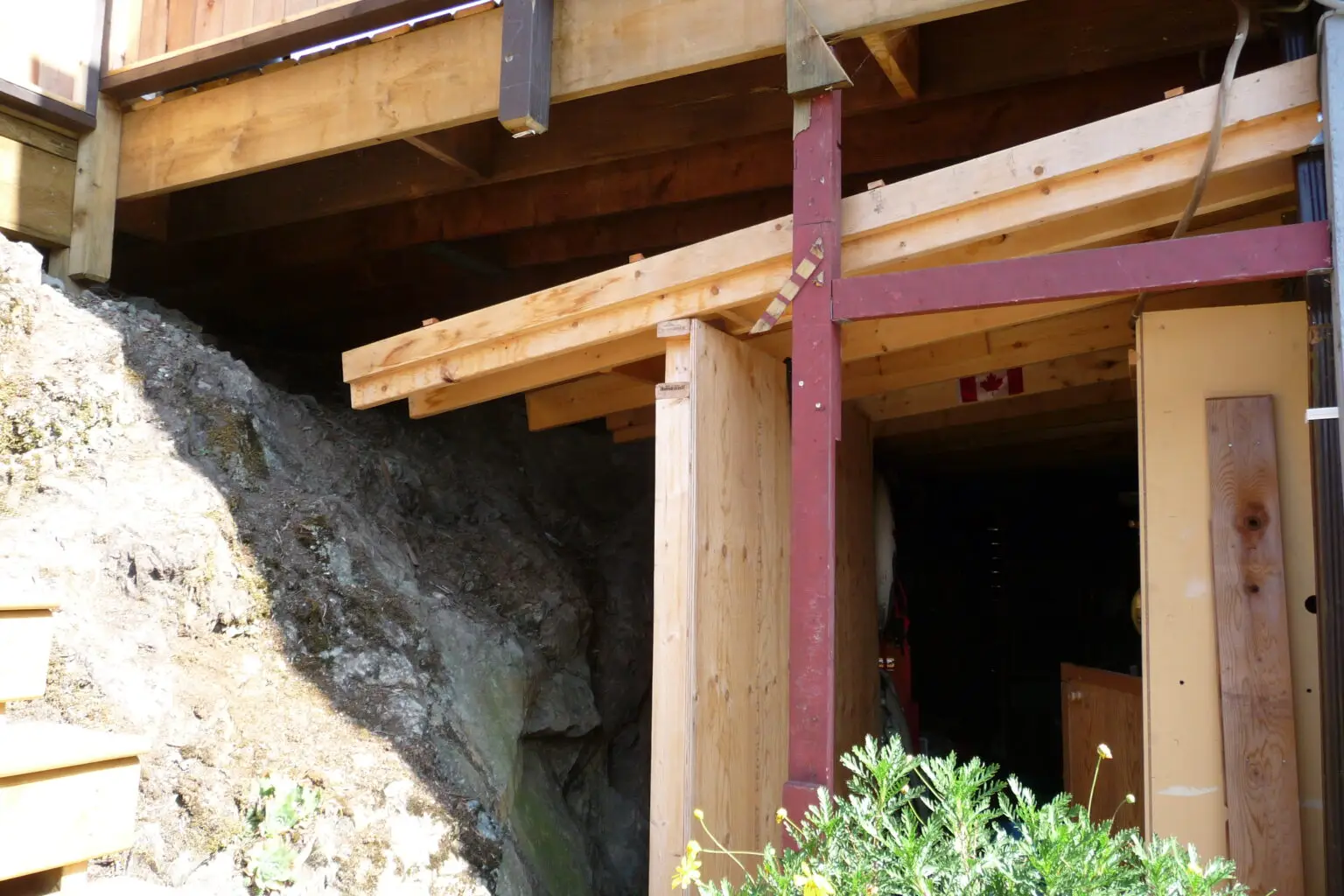 Before photo showing the under-deck storage area with an unfinished appearance, including exposed wooden beams and a tarp-covered entrance. The bare rock face next to the storage space is visible, with no greenery or landscaping.