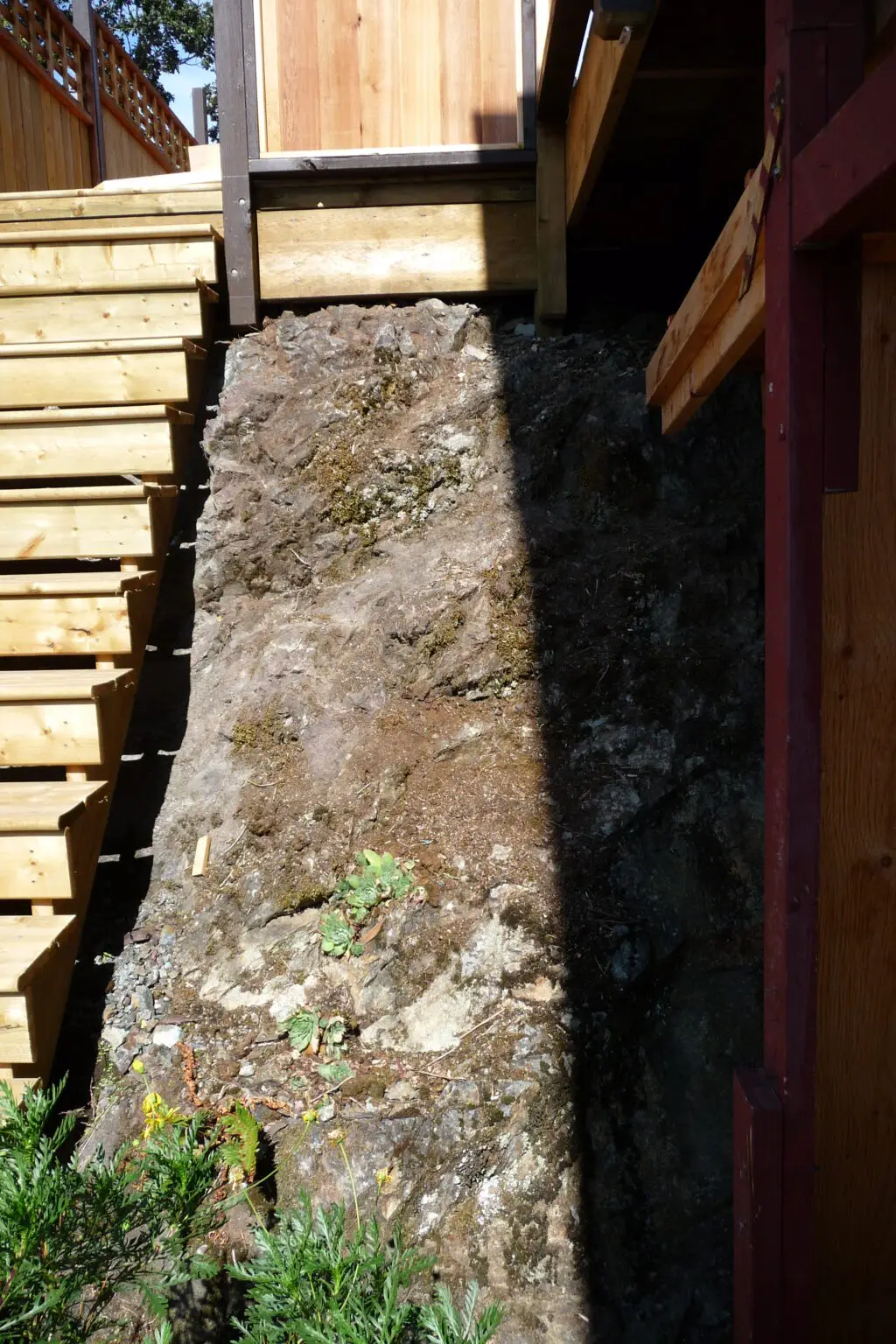 Photo showing the bare, exposed rock face next to newly installed deck stairs. The rocky surface highlights the challenge of landscaping in the area, with minimal existing greenery and difficult terrain.