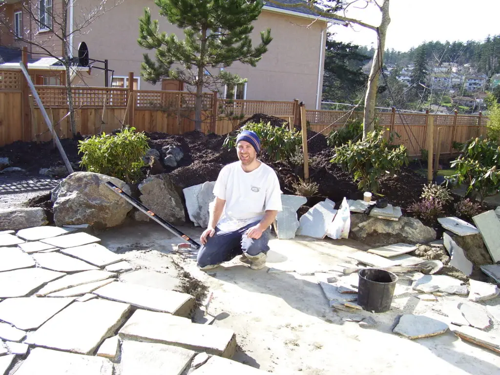 A landscaper kneeling on a patio under construction, surrounded by natural stone pieces and tools, with a background of shrubs, trees, and wooden fencing in a residential garden setting. The image captures the progress of a landscaping project in 2007