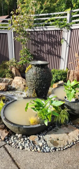 An urn fountain integrated into a multi-tiered water feature, where the water spills from the urn into the surrounding fountain landscape. One bowl overflows delicately into another, and in another section, water streams naturally into a crafted stream, mimicking a natural brook. This setup, surrounded by rich plant life and pebble accents, creates a dynamic and organic water display within the garden.