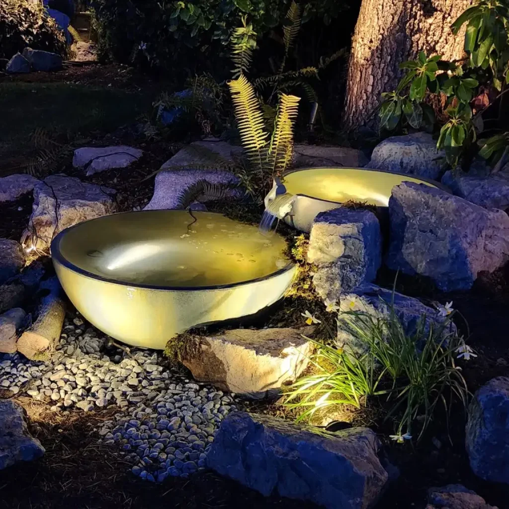 A serene garden water feature illuminated at night, featuring two cascading bowls with gently flowing water. The bowls are surrounded by large rocks, adding to the natural feel. Soft lighting highlights the water and the surrounding foliage, including ferns and small flowering plants. The backdrop consists of a tree trunk and lush greenery, creating a peaceful and ambient night-time garden scene.