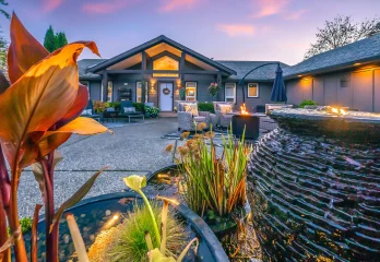 Stunning outdoor living space at sunset featuring a tranquil water fountain surrounded by lush aquatic plants in the foreground. The scene transitions to a welcoming patio with cozy wicker furniture and a fire table, all framed by a modern home with warm outdoor lighting and vibrant landscaping